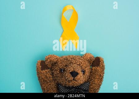 Top view of yellow ribbon with brown teddy bear on blue background, international childhood cancer day concept Stock Photo