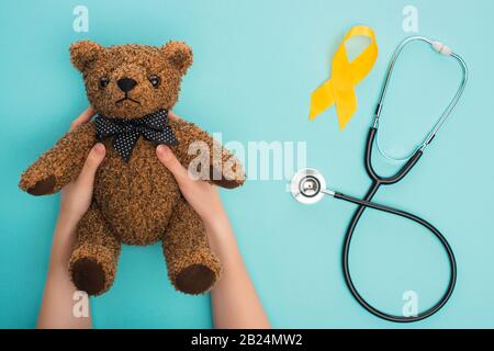 Cropped view of woman holding teddy bear near yellow awareness ribbon and stethoscope on blue background, international childhood cancer day concept Stock Photo