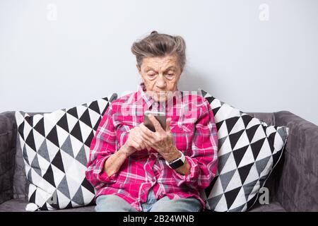 Theme old person uses technology. old gray-haired Caucasian woman with wrinkles sits home in living room on sofa and uses mobile phone in hands, looks Stock Photo