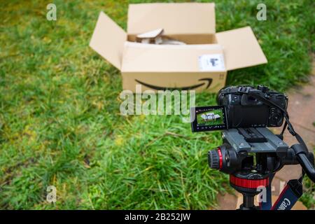 Paris, France - Feb 23, 2020: Vlogger setup for unboxing process from Amazon Prime e-commerce retailer with Panasonic GH5 mirrorless camera on tripod and cardboard box with logotype in grass Stock Photo