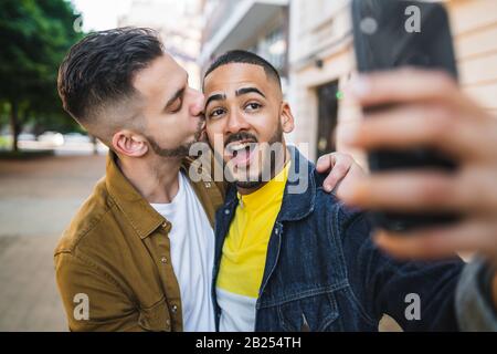 Portrait of happy gay couple spending time together and taking a selfie with mobile phone in the street. Lgbt and love concept. Stock Photo
