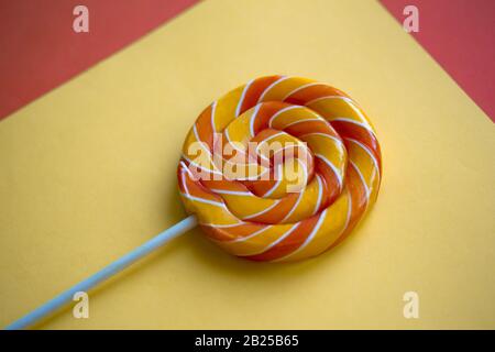 Bright swirl lollipop on yellow and orange background. Festive background with colorful twisted candy. Close up of rainbow round sweet on stick. Stock Photo