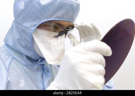 An engineer working in a clear room wearing a special uniform and protective glasses holds silicon wafer with microchips in hands in white gloves and Stock Photo