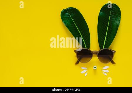 Rabbit face made of green leaves and flowers with sunglasses on yellow background. Flat lay minimal concept for Easter. Stock Photo