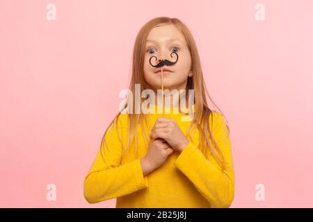 Portrait of cute little ginger girl holding funny paper mustaches and looking at camera with surprised eyes, having fun, wearing masquerade accessory. Stock Photo