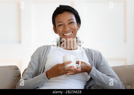 Portrait of happy biracial woman relax on couch drinking coffee Stock Photo