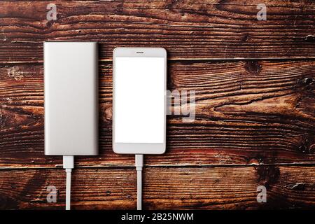 A white Powerbank charges a smartphone on a wooden background. Universal external battery for gadgets Free space and minimalistic composition. Stock Photo
