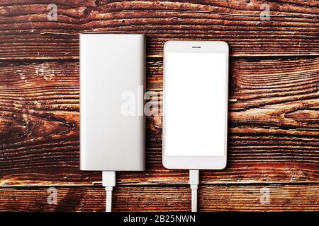 A white Powerbank charges a smartphone on a wooden background. Universal external battery for gadgets Free space and minimalistic composition. Stock Photo