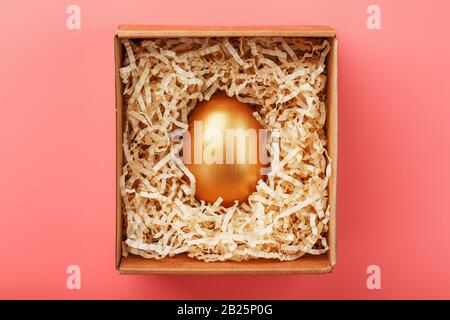 Golden egg in a cardboard box with shavings on a pink background. The concept of exclusivity, a special surprise, an expensive gift. The concept of mi Stock Photo