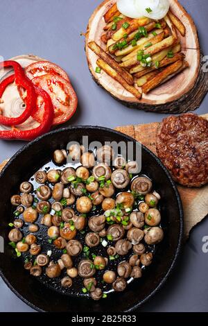 toasted golden champignon mushrooms sprinkled with green onions in a cast iron pan. juicy cutlet with french fries and vegetables on forest wooden coa Stock Photo