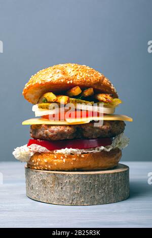 delicious burger with a large meat cutlet with vegetables, cheese and fries on a wooden forest stand on a gray background closeup. Stock Photo