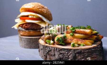 fried potatoes with mushrooms and a juicy burger on forest wooden boards. home fast food. Stock Photo