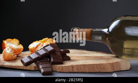bottle of white wine with pieces of milk chocolate and tangerine on a wooden forest stand on a dark background. romantic evening dinner for two close-