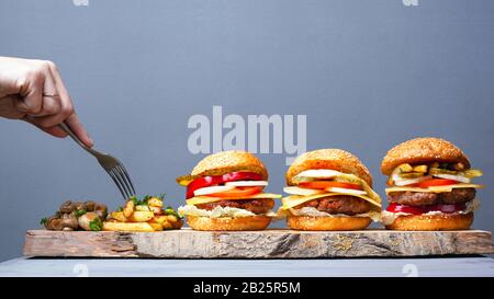 delicious rustic fast food cuisine. juicy 3 burgers potatoes and mushrooms on a gray background. eat with a fork. Stock Photo