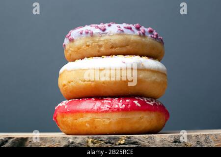 three donuts with colored glaze of violet red and white with powder stand on top of each other on a   stand on a gray background closeup. Stock Photo
