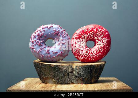 two round glazed donuts sprinkled with sweet icing red and purple on a wooden base on gray background. sweet bun on a decorative forest stand. Stock Photo