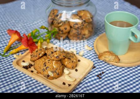 chocolate chip and nuts cookies decorated on a wooden plate with a cup of coffee, a cookie jar and flowers on a blue and white checked table cloth Stock Photo