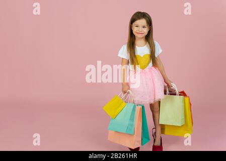Girl with many shopping bags on pastel pink backgound. Lovely sweet moments of little princess, pretty friendly child having fun to camera. Stock Photo