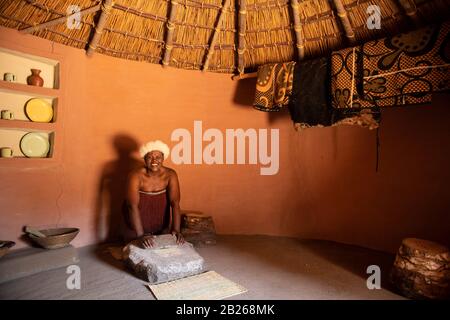 Basotho woman grinding maize in a traditional hut, Thaba Bosiu Cultural Village, Lesotho Stock Photo