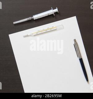 Template of white paper with pen, thermometer and syringe for injection on dark wenge color wooden background. The provision of medical and pharmacological services. With empty space for text. Stock Photo