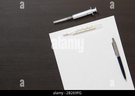 Template of white paper with pen, thermometer and syringe for injection on dark wenge color wooden background. The provision of medical and pharmacological services. With empty space for text. Stock Photo