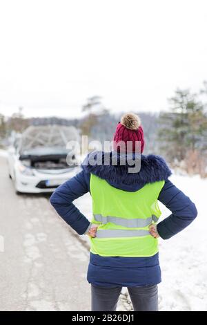 Rear view of stressed woman in reflective vest and broken car with opened hood in the background. Road trip problems and assistance concepts. Stock Photo