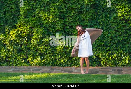 Happy stylish women hipster boho style dancing near green leaves wall building in sunny city, summer travel, nature love concept. Stock Photo