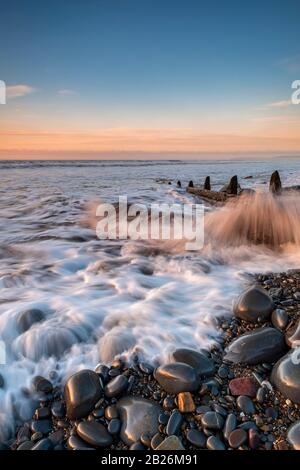 Incoming tide splashing around the pebbles and wooden sea defences gabions, on Westward Ho! Beach with sunset on the horizon, North Devon, South West, Stock Photo