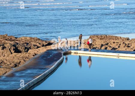 People swimming in the rock pool at Westward Ho!, North Devon, UK surrounded by rocks with blue seas and the reflection of swimmers in the water Stock Photo