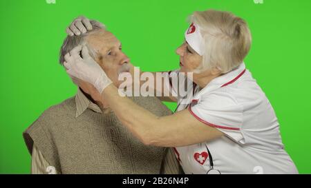 Kind mature woman nurse caregiver supporting to old elder man help with problem, female doctor therapist in uniform examines man and give treatment tips to senior patient Stock Photo