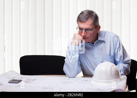 Senior pensive architect of engineer at a table with blueprint and white hard hat in a office Stock Photo