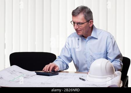 Senior architect or engineer calculating a project in an white office with blueprint, hard hat und calculator on a table Stock Photo