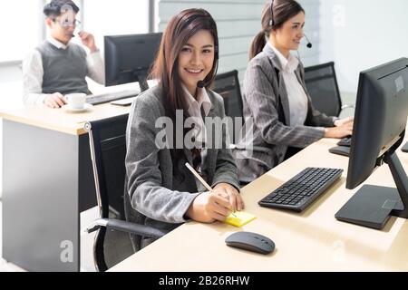 Young adult friendly and confidence operator woman agent smiling with headsets working in call centre with her colleague team working. Stock Photo
