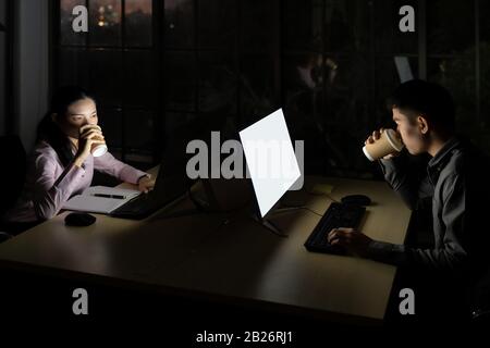 Young adult asian businessman and woman drinking hot cofee while working late at night in their office with desktop computer and laptop. Using as hard Stock Photo