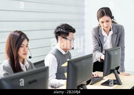 Young adult confidence with smiling female supervisor standing in front of working table of operator team ain a call centre to monitor her employee. U Stock Photo