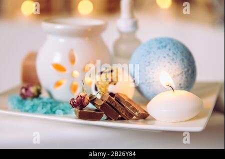 Home spa products on wooden white ceramic tray: bath bomb, aroma bath salt, aroma oil lamp, essential and massage oils, pieces of chocolate. Stock Photo