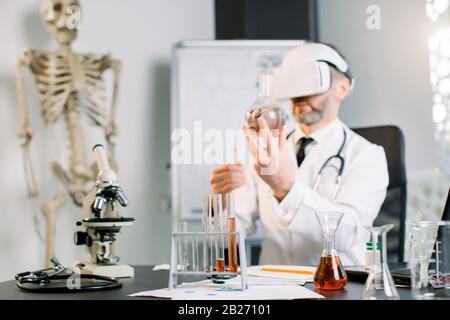 close up of man scientist wearing vr goggles and holding flask, making test in clinical laboratory. Doctor scientist wearing virtual reality glasses Stock Photo