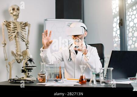 Caucasian man lab worker wearing VR goggles at work. Science technician in virtual reality glasses working on virtual screen in laboratory. Science Stock Photo