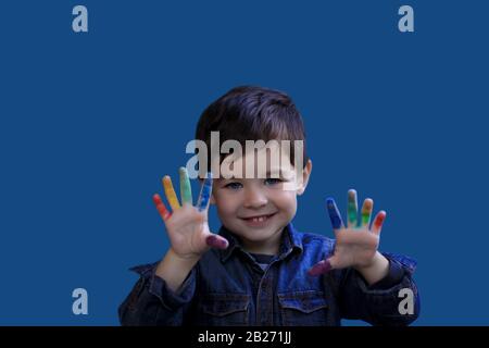 Cute little brunette boy with blue eyes, stands on an isolated blue background, shows fingers stained in colorful paint. Children's creativity. Rainbo Stock Photo