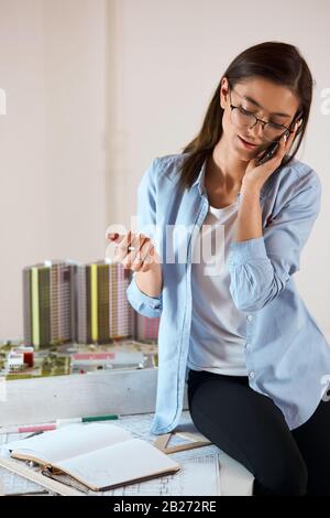 young clever designer talking with customers while sitting on the edge of the desk, woman consulting customers , vertical shot. close up photo Stock Photo
