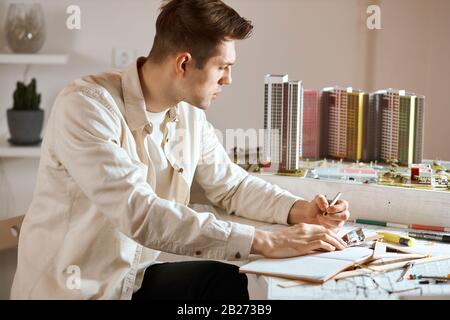serious attractive guy drawing a new building on blueprint, looking at the small model of house and thinking about construction. close up side view ph Stock Photo