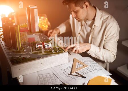 hardworking blonde architect late night working at home, measuring a model with a ruler, sitting at the table, close up side view photo. Stock Photo