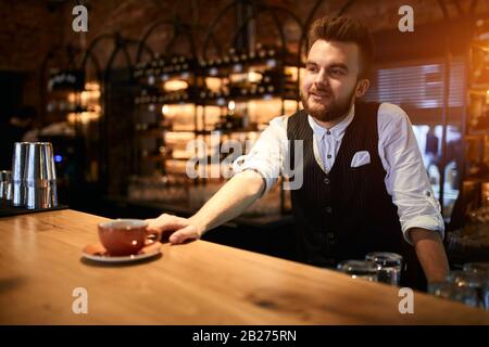 Portrait of smiling bearded stylish manstanding at table while having job in confectionary shop, offering a cup of tea. beverage Occupation concept. C Stock Photo