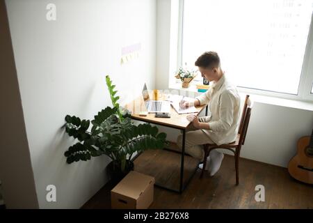 Concentrated serious hardworking young blonde man using laptop and taking notes at home in the living room, guy writing resume, side view photo. Stock Photo