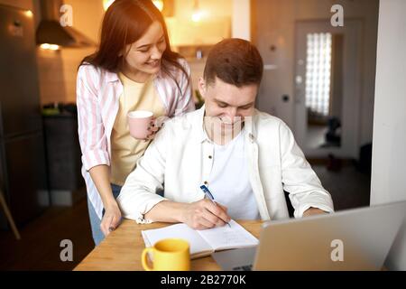 happy man making notes, his wife standing behind him and smiling, family business, close up photo, interest, hobby, education online Stock Photo