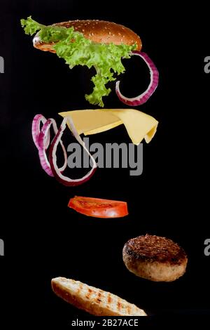 Flying ingredients burger or cheeseburger on a small wooden cutting board isolated on a dark background. Burger floating in the air above the table Stock Photo