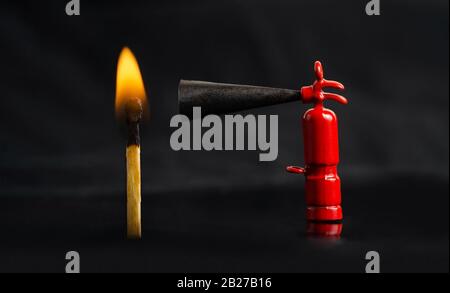 Tiny fire extinguisher close to a burning match against a lit black background, macro shot Stock Photo