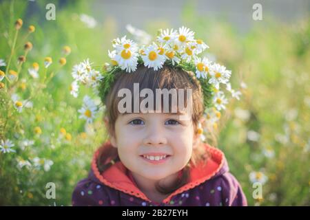 A little girl in a wreath of daisies smiles and sits in the green grass in summer. Sunny day. Stock Photo