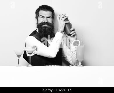 handsome bearded barman with long beard and mustache has stylish hair on funny face holding shaker and made alcoholic cocktail, in vintage suede leather waistcoat on white studio background Stock Photo