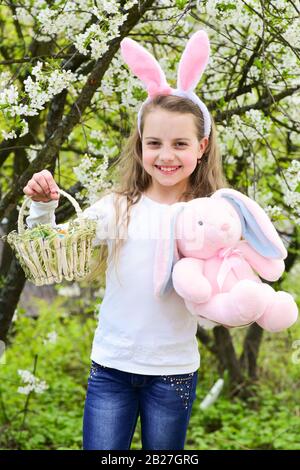 Happy girl smiling in bunny ears, rabbit and basket. Child playing in garden with blossoming trees. Childhood, youth and growth. Easter and spring concept. Holidays celebration with presents and toys. Stock Photo